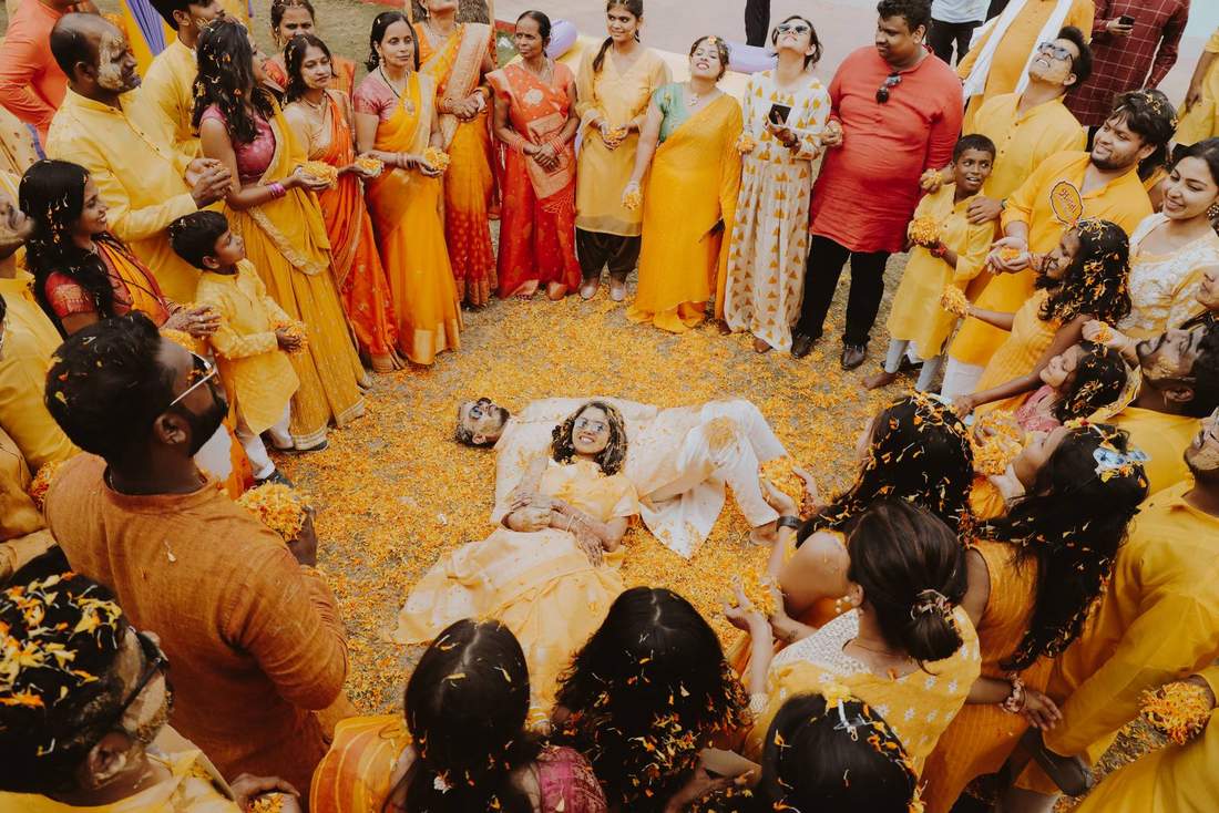 Overhead shot of a couple lying amid flower petals during haldi, encircled by family